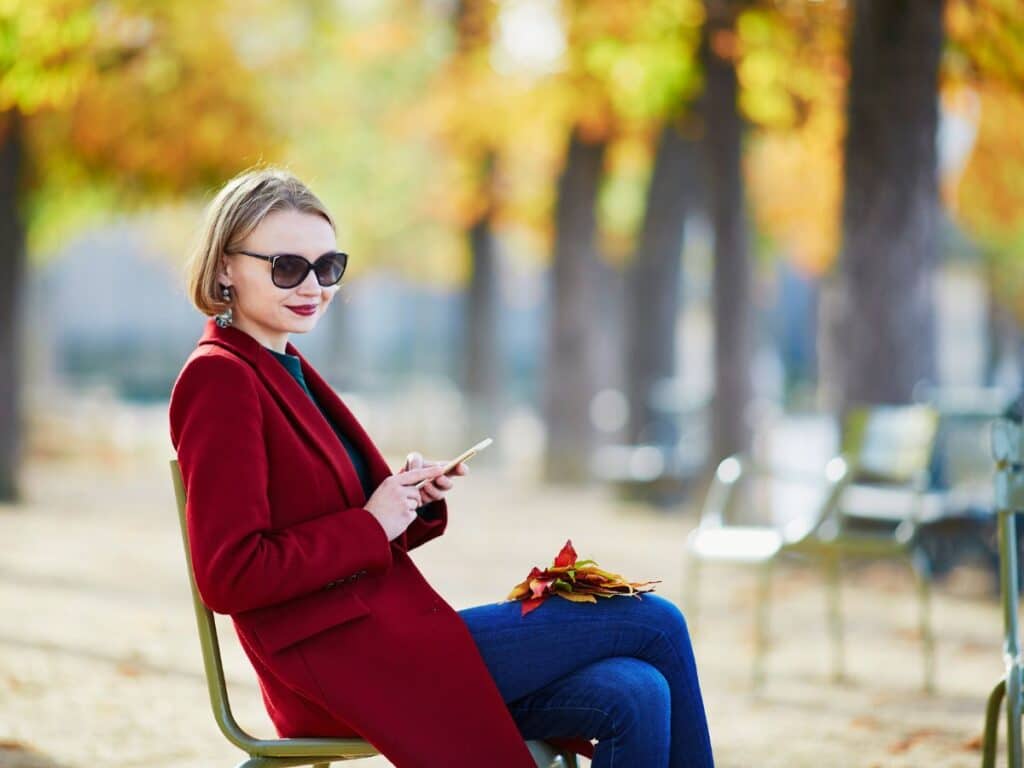 A woman in a deep red coat sits in a park with autumn leaves, holding her phone and wearing oversized sunglasses, embodying relaxed Parisian chic with a bold twist.