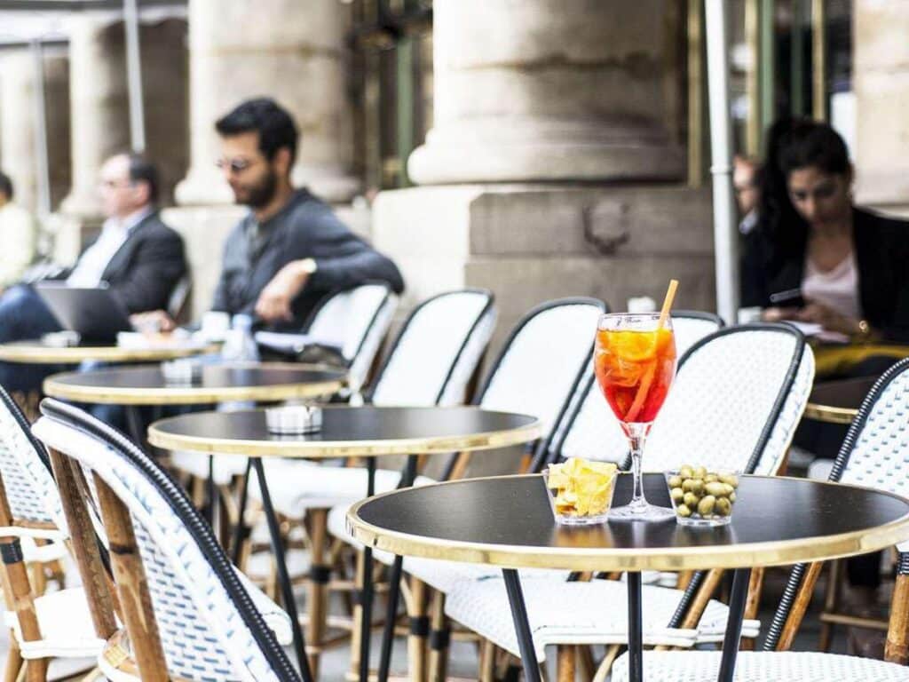 Glass of orange aperitif with chips and olives on a café table, surrounded by white bistro chairs; blurred background shows casually dressed people working or relaxing, showcasing typical Paris café etiquette of savoring time and space.
