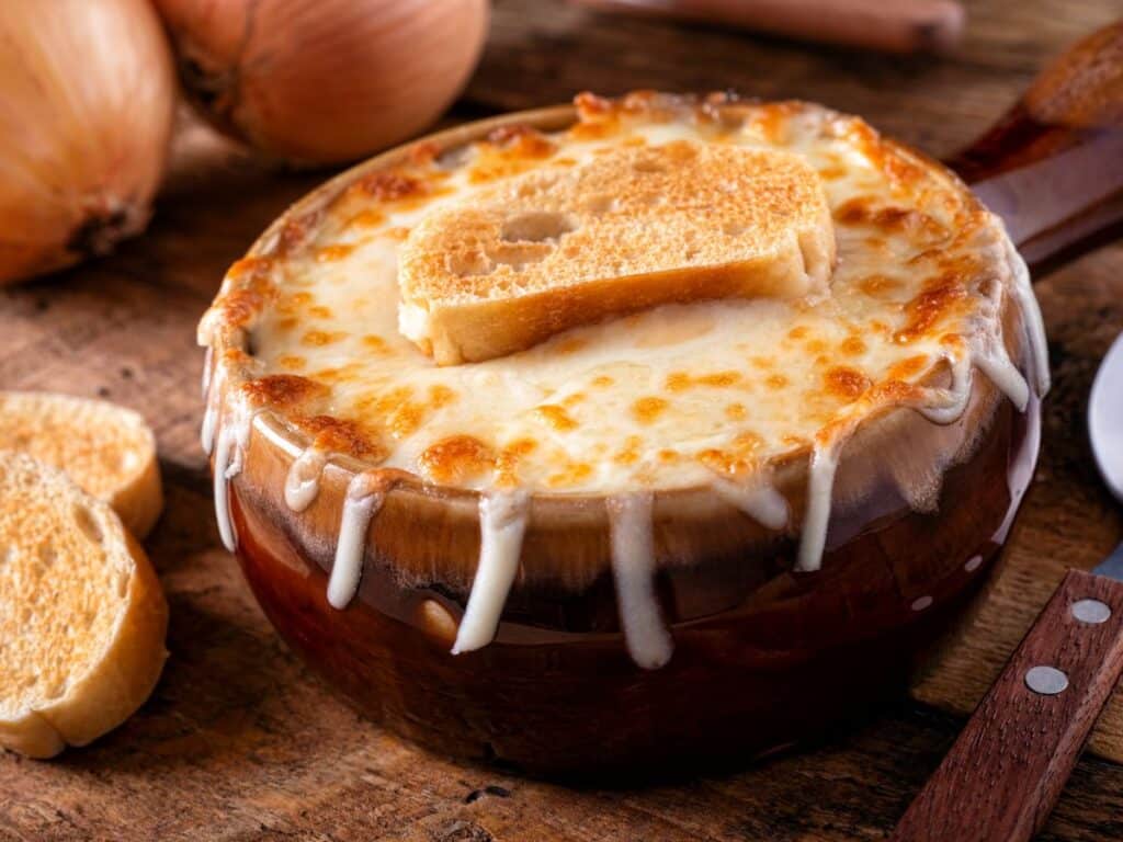 Rustic ceramic bowl of French onion soup with thick melted cheese dripping down the sides and a crispy baguette slice on top, surrounded by raw onions and bread.