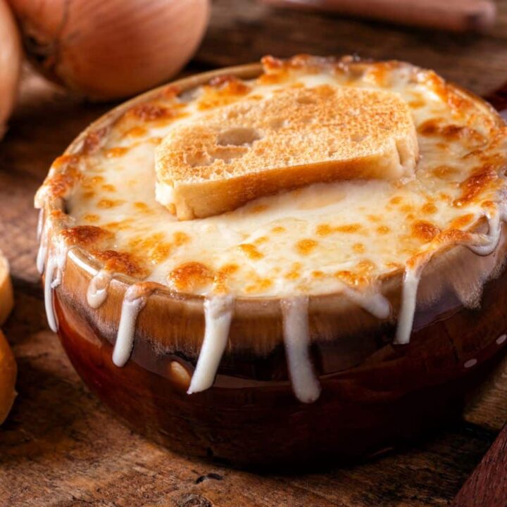 Rustic ceramic bowl of French onion soup with thick melted cheese dripping down the sides and a crispy baguette slice on top, surrounded by raw onions and bread.