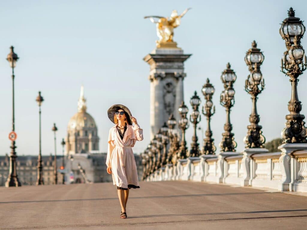 A woman walks across the ornate Pont Alexandre III bridge in Paris, wearing a flowing pale dress and a wide-brimmed black hat—an iconic scene radiating Parisian chic glamour.