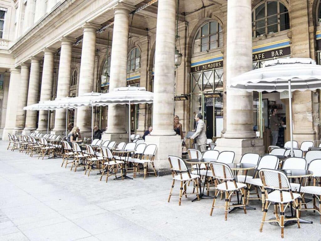 Expansive Parisian café terrace outside a neoclassical building with tall columns and striped umbrellas, featuring orderly rows of black tables and rattan chairs — a prime example of French café etiquette emphasizing elegance and spatial harmony.