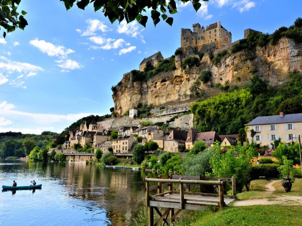 Scenic view of Beynac-et-Cazenac in the Dordogne region of France, with stone houses lining the riverbank and a historic castle perched on a limestone cliff above. A small canoe floats on the peaceful water under a bright blue sky.