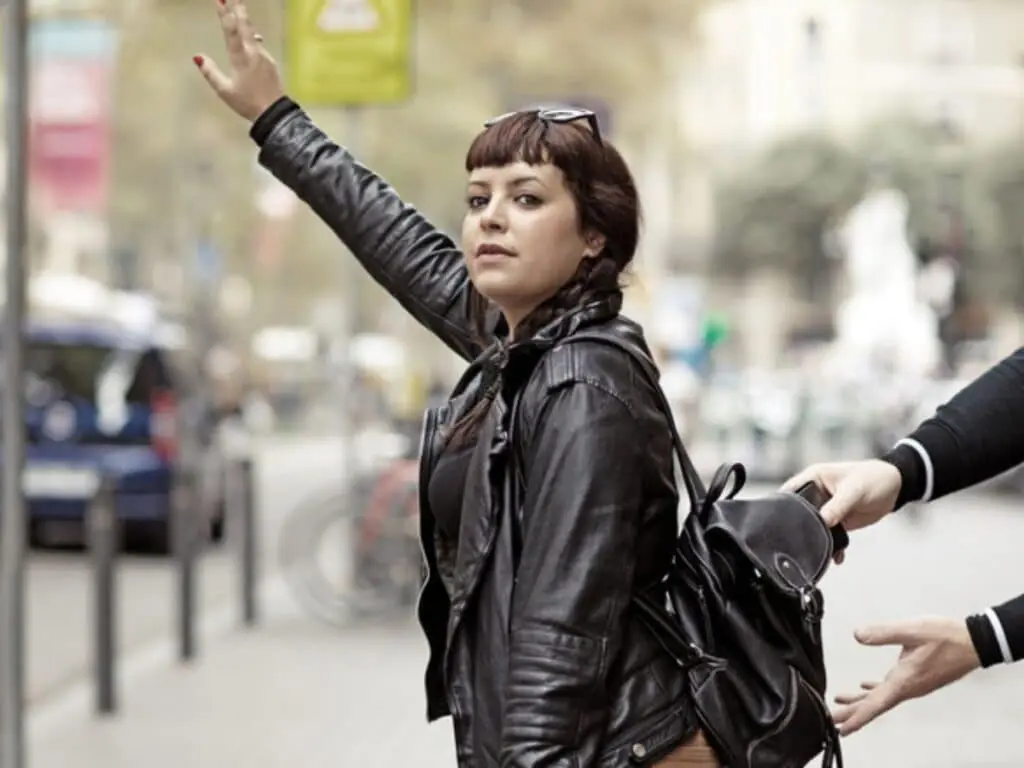 A woman raising her hand on a city street, unaware that someone behind her is stealing from her backpack. This common Paris scam targets distracted tourists in public areas.