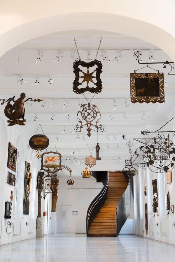 Ornate wrought iron shop signs hang from the ceiling in a bright gallery at the Musée Carnavalet, showcasing Parisian commercial history with intricate metalwork and a winding wooden staircase at the far end.