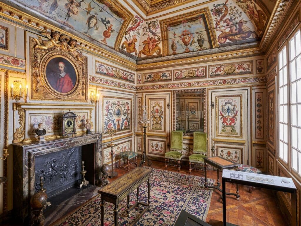 Lavishly decorated 17th-century room inside the Musée Carnavalet featuring a marble fireplace, green velvet chairs, and richly painted walls and ceilings with baroque motifs and a portrait of Cardinal Richelieu.