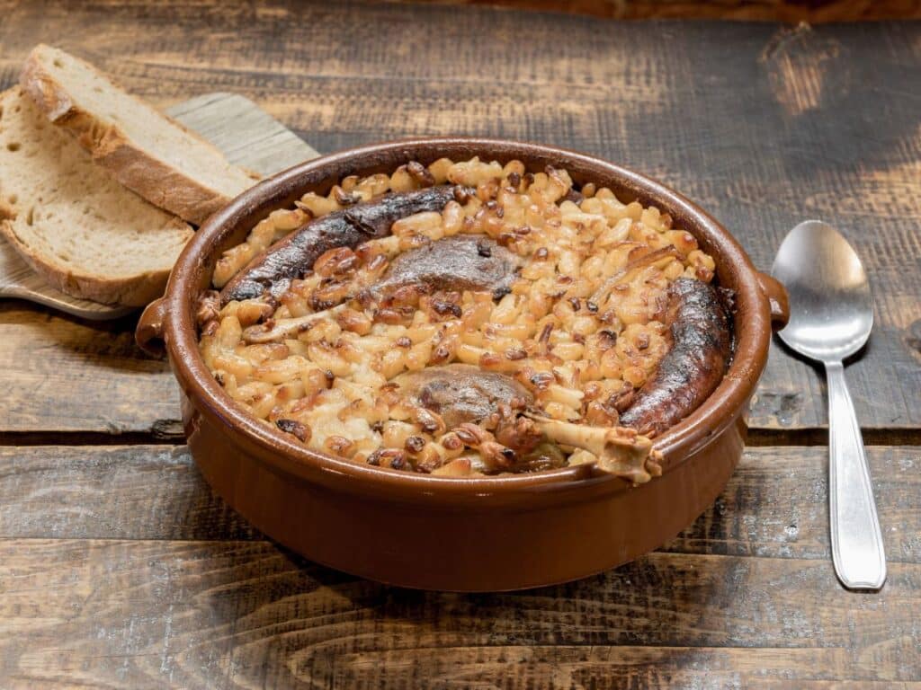 Hearty cassoulet filled with white beans, duck confit, and browned sausages in an earthenware pot, served with thick slices of crusty country bread on a wooden table.
