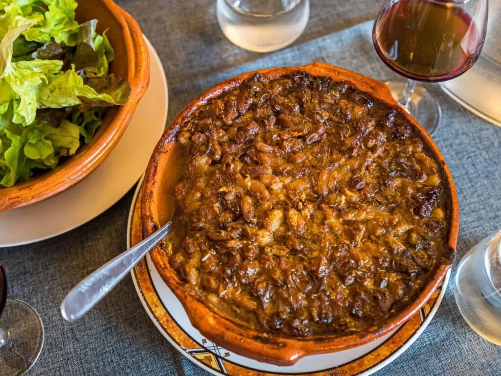 A traditional French cassoulet served in a rustic clay dish, featuring a rich, caramelized crust of slow-cooked beans and meats, paired with a side salad and red wine on a linen-covered table.