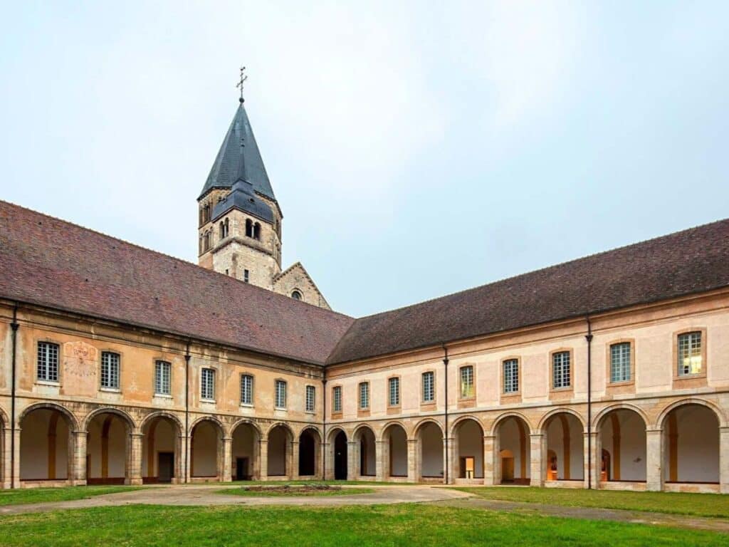 The courtyard of Cluny Abbey with arched walkways and a central grassy lawn, backed by a Romanesque church tower—once the largest church in medieval Europe.