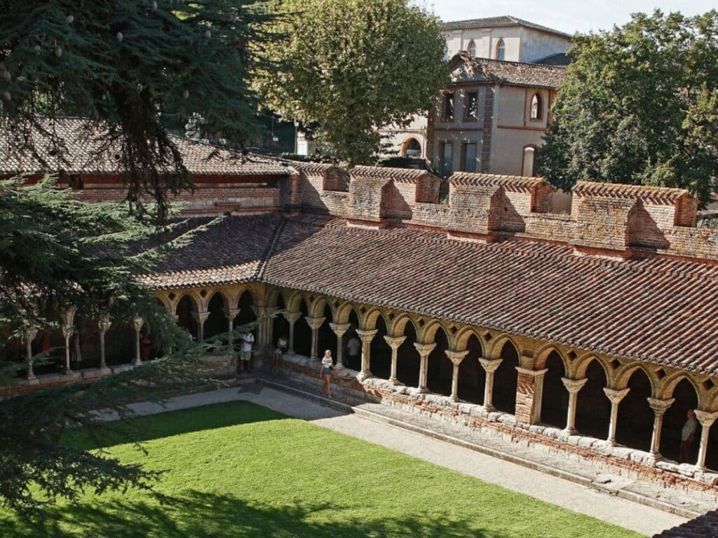 A red-brick cloister with paired columns and a central lawn at Moissac Abbey in southwestern France, with visitors walking through its arcaded galleries.