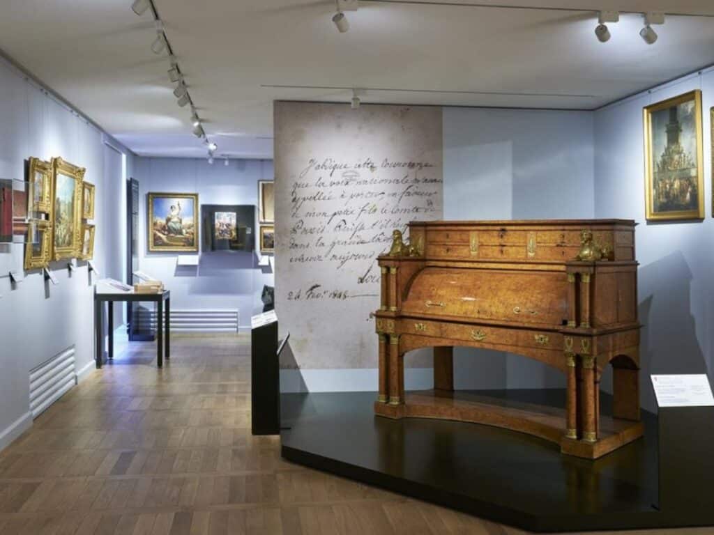 Museum gallery at the Musée Carnavalet displaying framed paintings, historic artifacts, and a polished wooden desk with neoclassical detailing, all under focused track lighting.