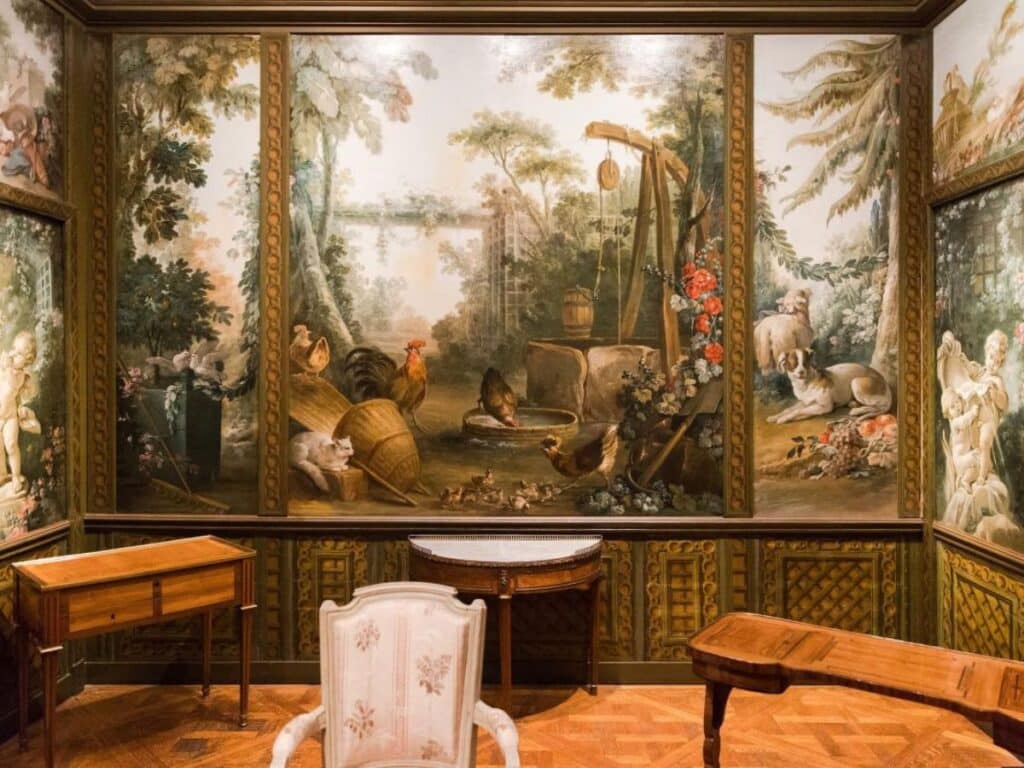 Pastoral-themed room at the Musée Carnavalet featuring scenic wall murals with roosters, dogs, and trees, along with antique wooden furniture and soft lighting that evokes a rustic charm.