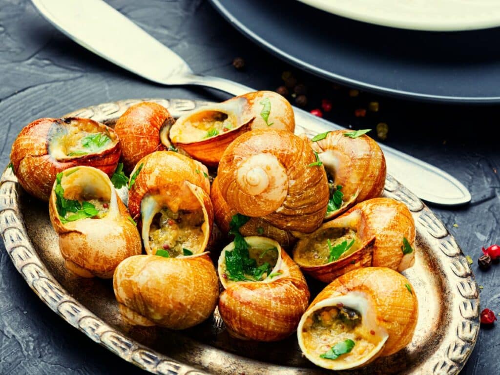 A silver platter of baked escargot stuffed with garlic butter and herbs, some shells slightly open to reveal the savory filling. This rich and traditional presentation visually supports the cultural reasons why the French eat snails.