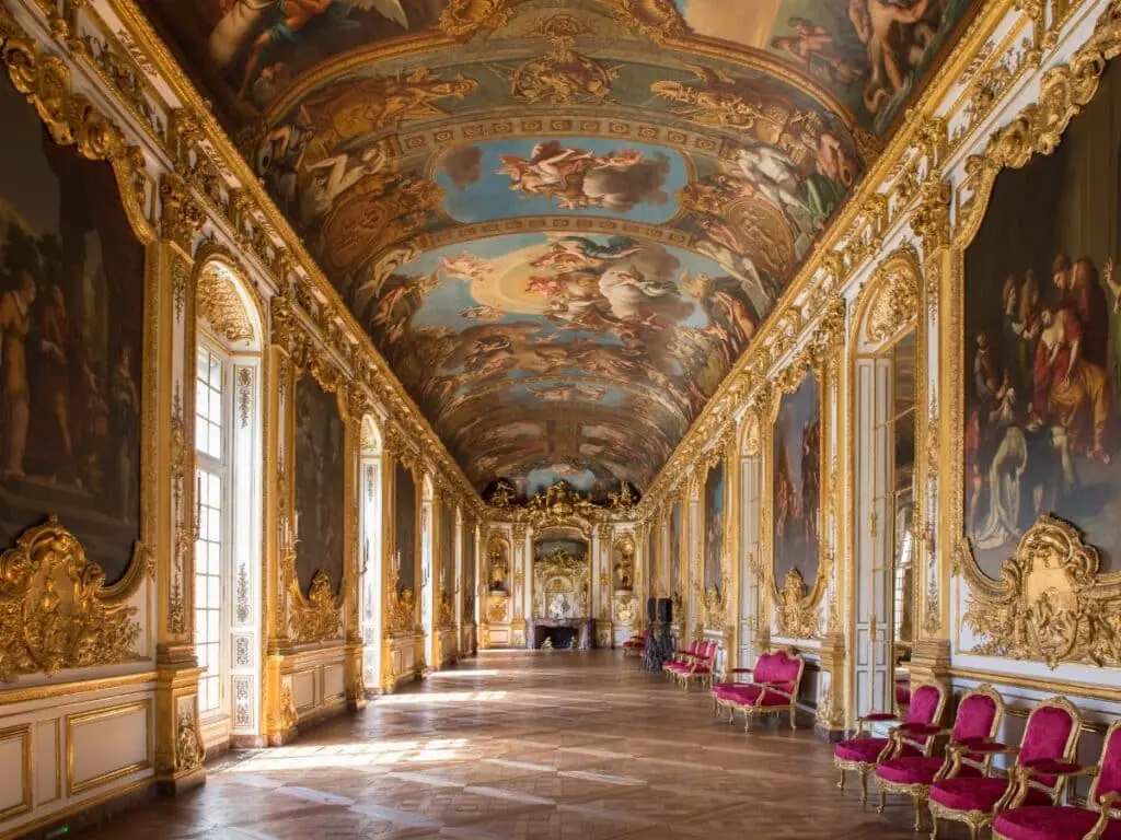 The grand gallery at the Musée Carnavalet with opulent gold-trimmed walls, crimson velvet chairs, and dramatic ceiling frescoes depicting mythological scenes, illuminated by natural light from tall windows.